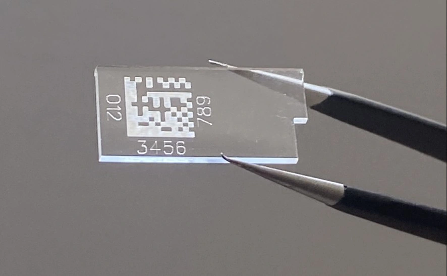Pièce transparente en CR39 tenue par une pince, avec un DataMatrix miniature et des caractères gravés